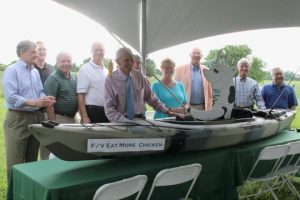 Chester County Economic Development Council President and CEO Gary W. Smith (front), a kayaking enthusiast, laughs as he receives a kayak with a cow at the helm to mark his 40-year tenure at CCEDC. Behind him (from left) are past and present CCEDC board chairs: Jerry Parker, Bill Bogle, Stan Schuck, Conrad Olie, Roger Huggins, Nancy Corson, John Snyder, Hanno Spranger, and Frank Krempa.